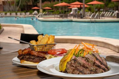 A plate with steak and a plate with a burger and fries on an outdoor table at Harry’s Poolside Bar & Grill, one of the beautiful scenic waterfront restaurants in Orlando. 