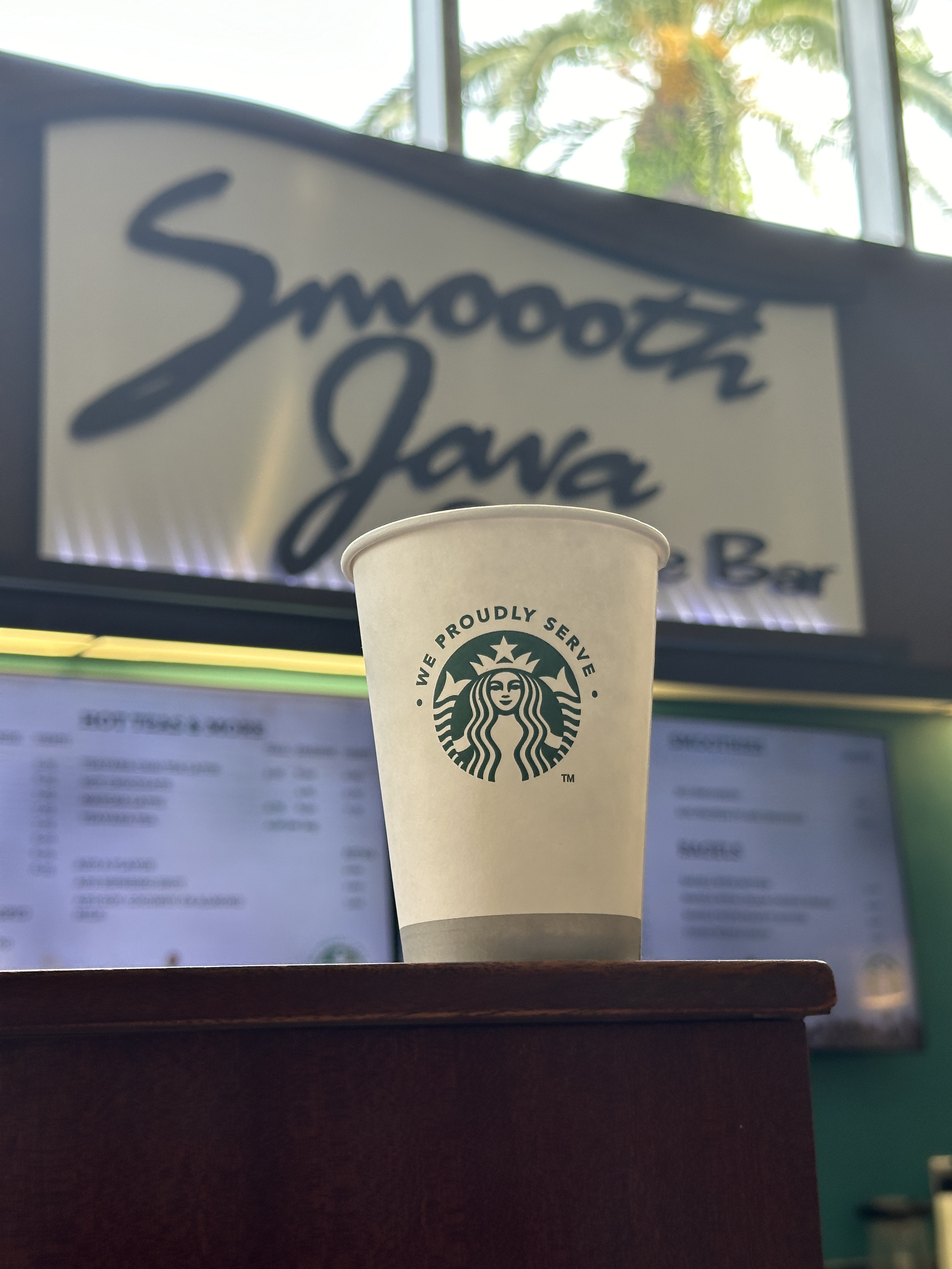 A white cup reading "We Proudly Serve" with the green Starbucks logo sits on a counter in front of a sign that reads Smoooth Java Bar. 