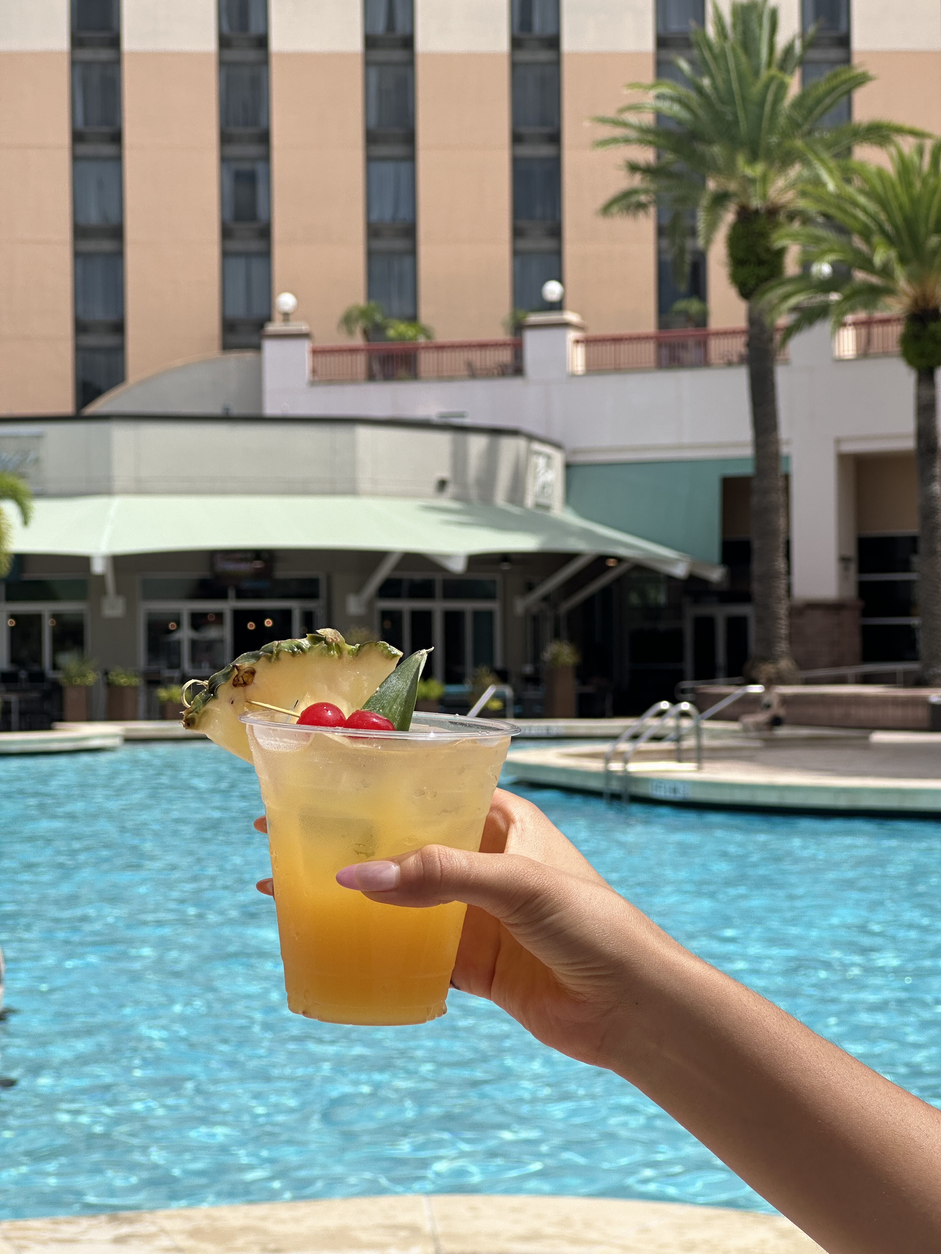 A woman's hand holds up a a tropical drink, with a pineapple wedge, in front of a pool and hotel. Get a drink at Harry's as one of several self-care practices that are hotel-friendly.