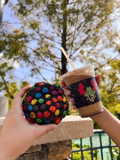 Two hands holding a chocolate cookie with colorful chocolate candies and an iced coffee in front of a background of trees.