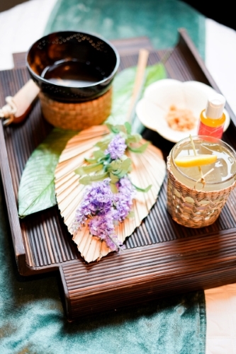 A wooden tray with a beverage, fan, and purple florals in a spa-like setting, ready for a spa treatment. 