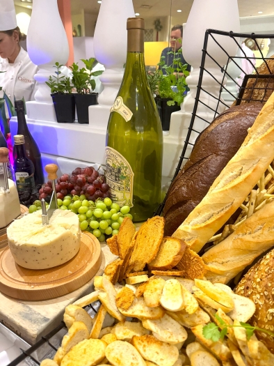 A table display with a wheel of cheese, a large empty wine bottle, red and white grapes, baguettes, and other sliced bread. 