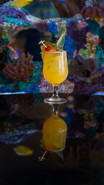 A yellow cocktail in a stemmed glass garnished with pineapple and a cherry sits on a dark reflective table in front of a glowing aquarium.