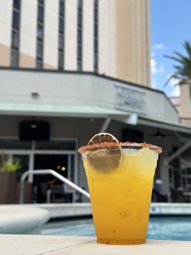  A cloudy orange cocktail with a dried citrus slice and orange sprinkled rim sits on the ground beside a blue pool with a tall building in the background. 