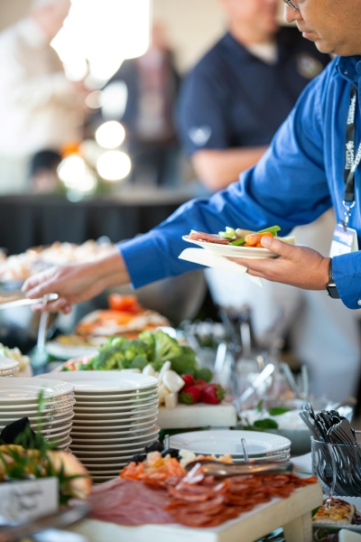 A man in a blue shirt with a lanyard helps himself from a buffet table with veggies and charcuterie. 