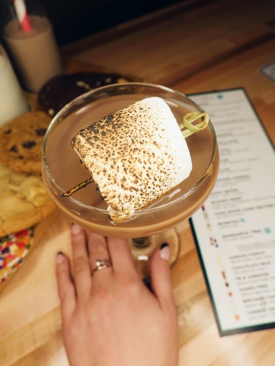 A hand holding the base of a martini glass with a milky brown liquid topped with an enormous toasted marshmallow on a skewer. Also on the table are a long white menu and a row of cookies. 