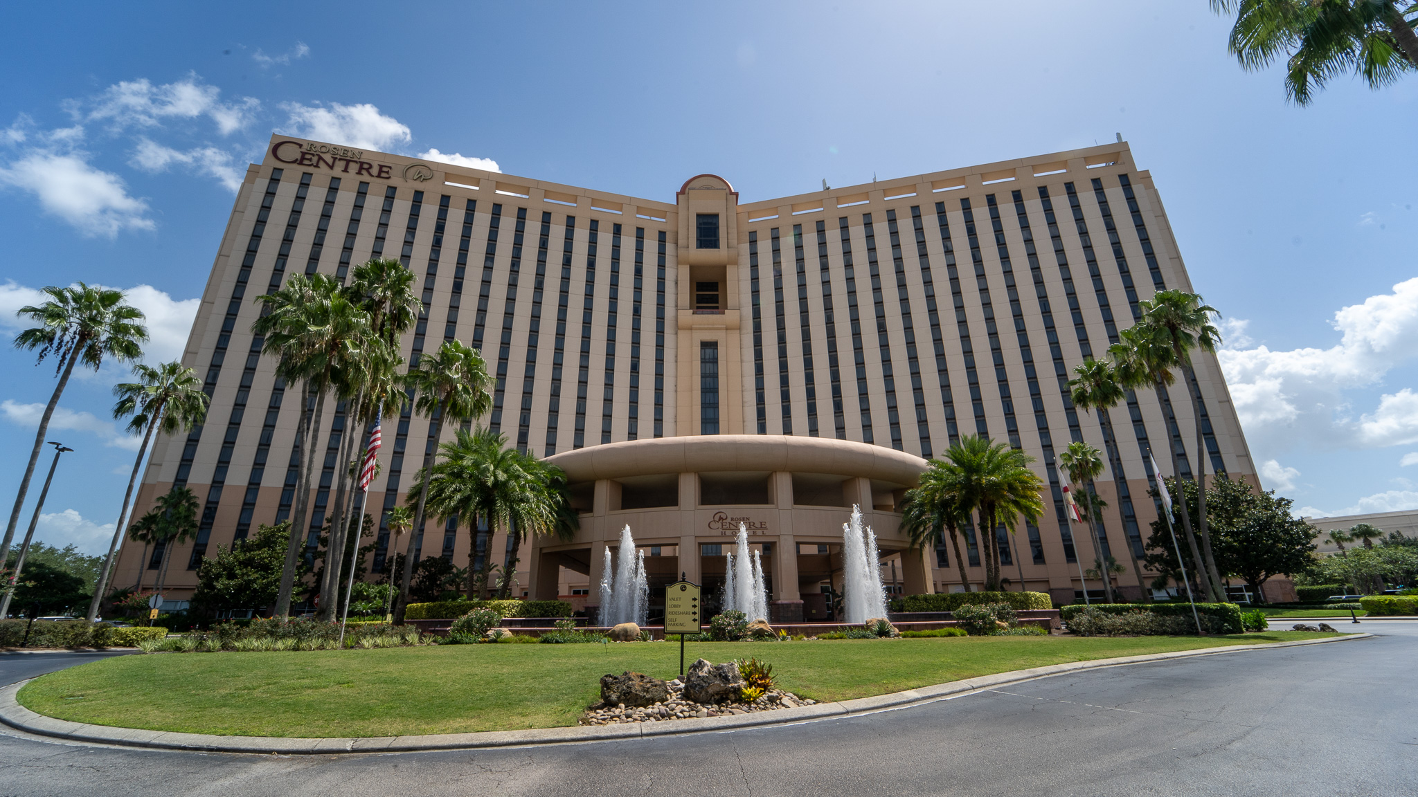 Beige facade of Rosen Centre hotel, featuring hundreds of hotel room windows, three water fountains spouting at the entrance and palm trees lining the front. Stay at Rosen Centre for your next OCCC event. 