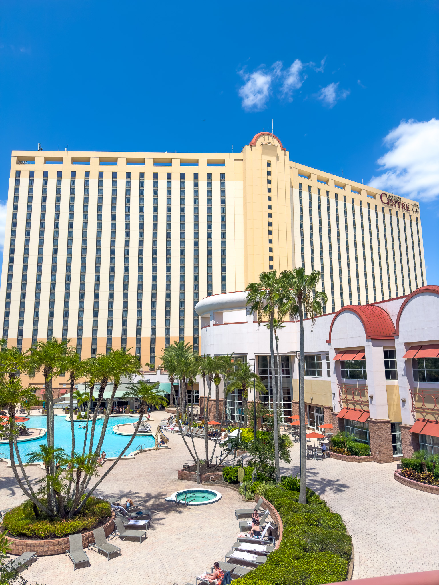 The exterior shot of the Rosen Centre hotel showing palm trees, a hot tub, pool chairs, adjacent buildings. 