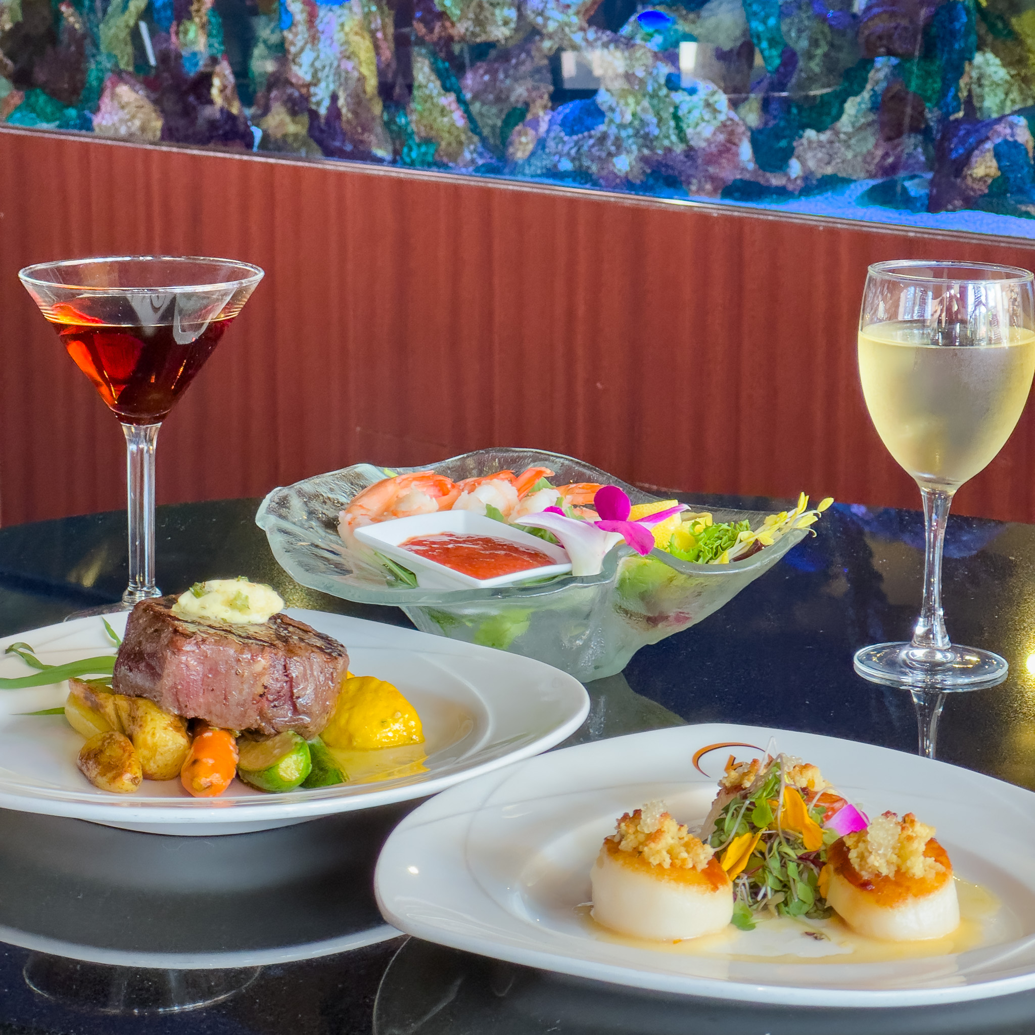 Three dishes of food, featuring meat, vegetables, and seafood, are on a table with two drinks at Everglades Restaurant at Rosen Centre.