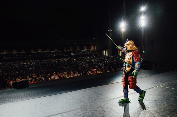 A performer in a sci-fi warrior costume stands on a bare stage in front of a dark crowd of hundreds of people. 