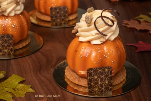 An orange pastry that resembles a pumpkin topped with icing and surrounded by wood grain and fall leaves. 