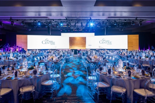 A large ballroom space with a grid of professional lights overhead, set with round tables and blue lighting. At the front of the room, two large screens project the Rosen Centre logo.