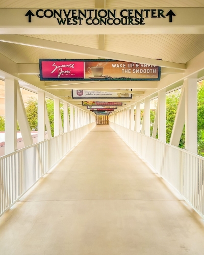 A covered outdoor walkway with white fencing and barricades surrounded by treetops leading to a set of doors in the distance.