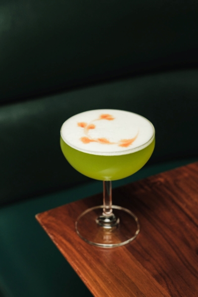 A lime green cocktail in a rounded martini glass topped with frothy egg whites and an orange floral design dusted on top. 