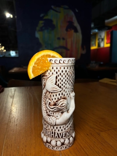 A white tiki cup with dark carvings of a mermaid garnished with an orange slice sitting on a wooden table in a dim room. 