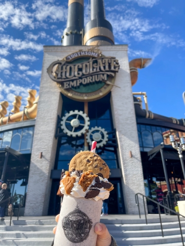 A hand holding a giant milkshake topped with a chocolate chip cookie in front of Toothsome Chocolate Emporium, the perfect place to satisfy your sweet tooth. 