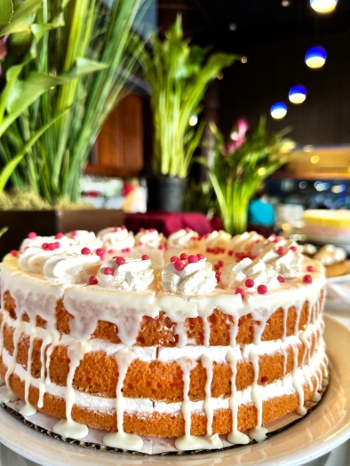 A beautiful three-tiered naked cake with white icing dripping down the side and red decorations on top sitting on a counter in front of tall green plants. 