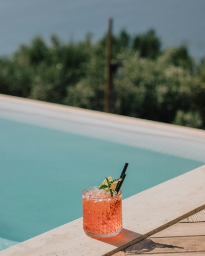 A pink cocktail in a tumbler sits on the edge of a pool overlooking green treetops.