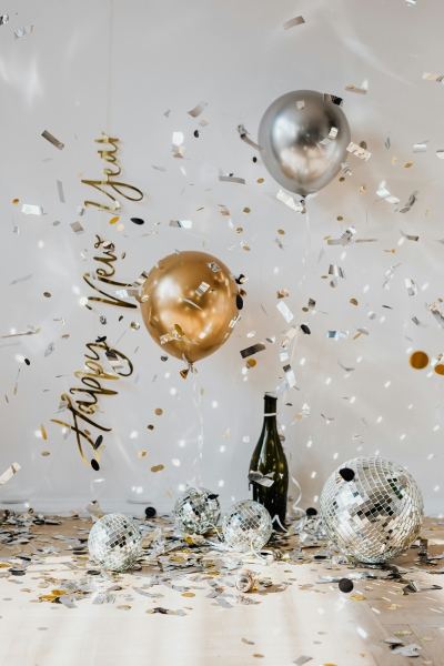 A bottle of Champagne sits amidst sparkling disco balls, confetti, a sign that reads “Happy New Year,” and a gold and silver balloon. 