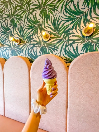 A hand holding an ice cream cone with swirled purple ice cream in front of a wall with arched cushions and floral wallpaper. 